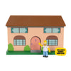 A Simpsons toy house set featuring a peach-colored two-story house with a brown roof, green bushes, and a small Homer Simpson figure standing in front. A yellow badge on the right reads 'INCLUDES HOMER SIMPSON!'.